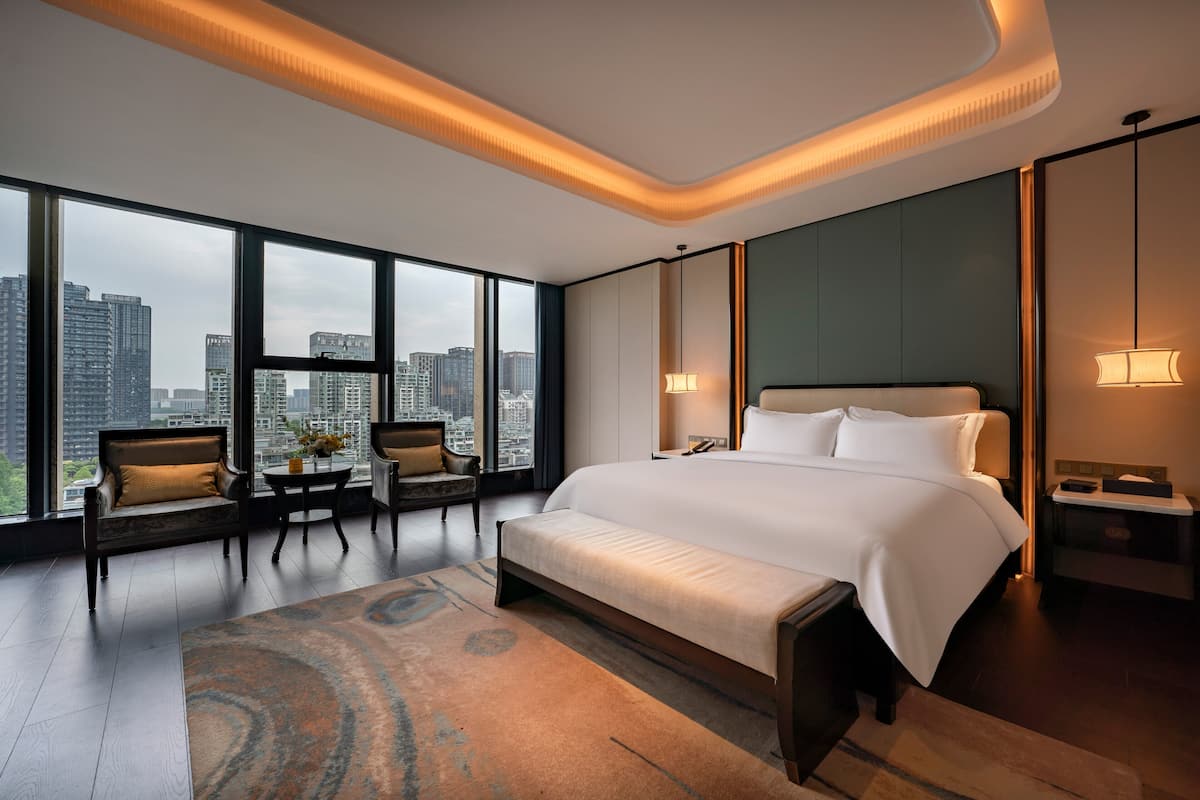 Une chambre d’hôtel moderne dotée d’un grand lit, d’un coin salon avec des fauteuils, d’une vue sur la ville et d’un panorama urbain visible à travers les fenêtres.