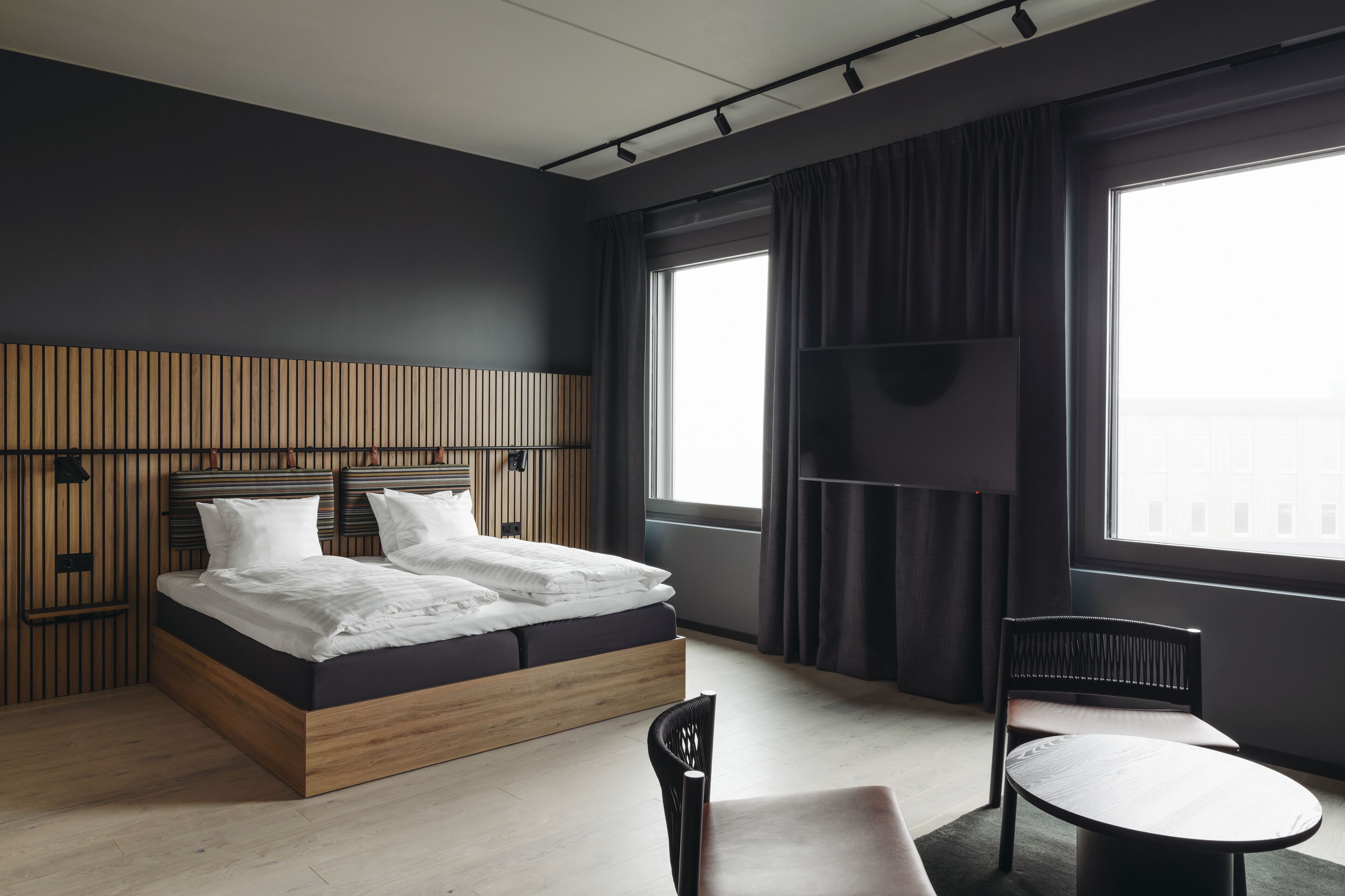 Una habitación de hotel moderna con cabecera de madera, una cama grande, televisor de pantalla plana, una mesa redonda y una silla.