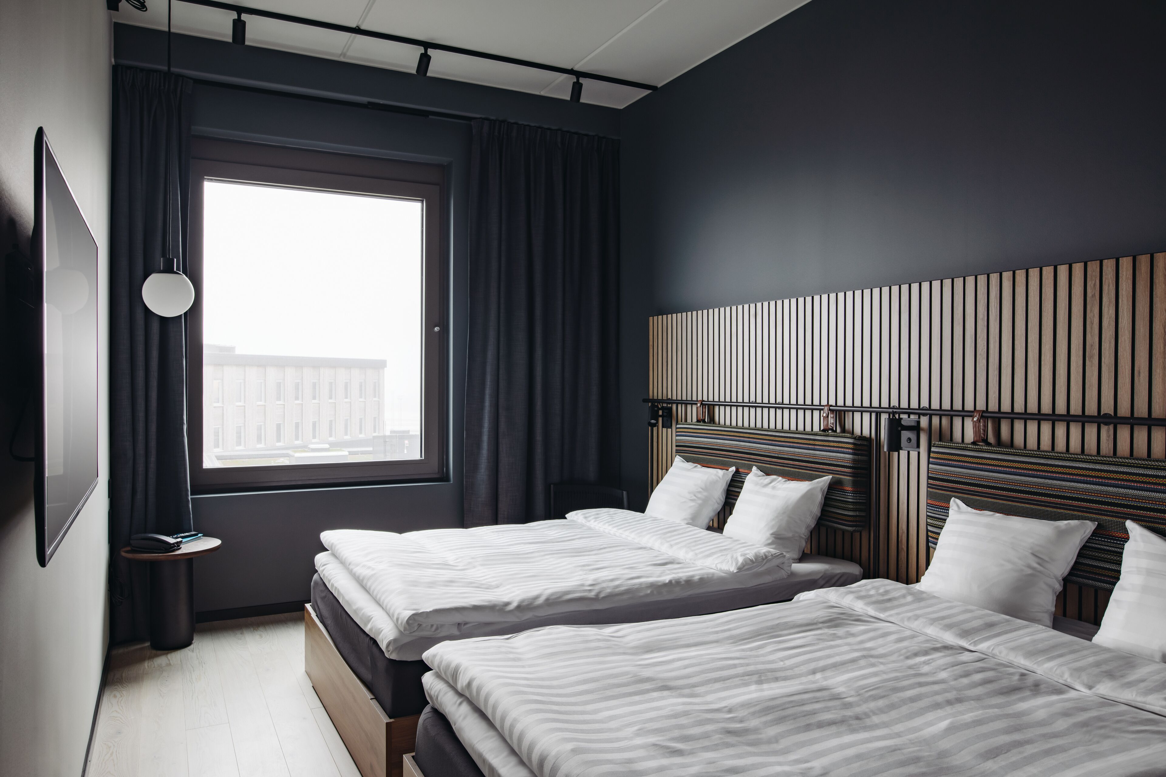Una habitación de hotel moderna con dos camas, un ventanal con cortinas, un televisor montado en la pared y un cabecero de madera.