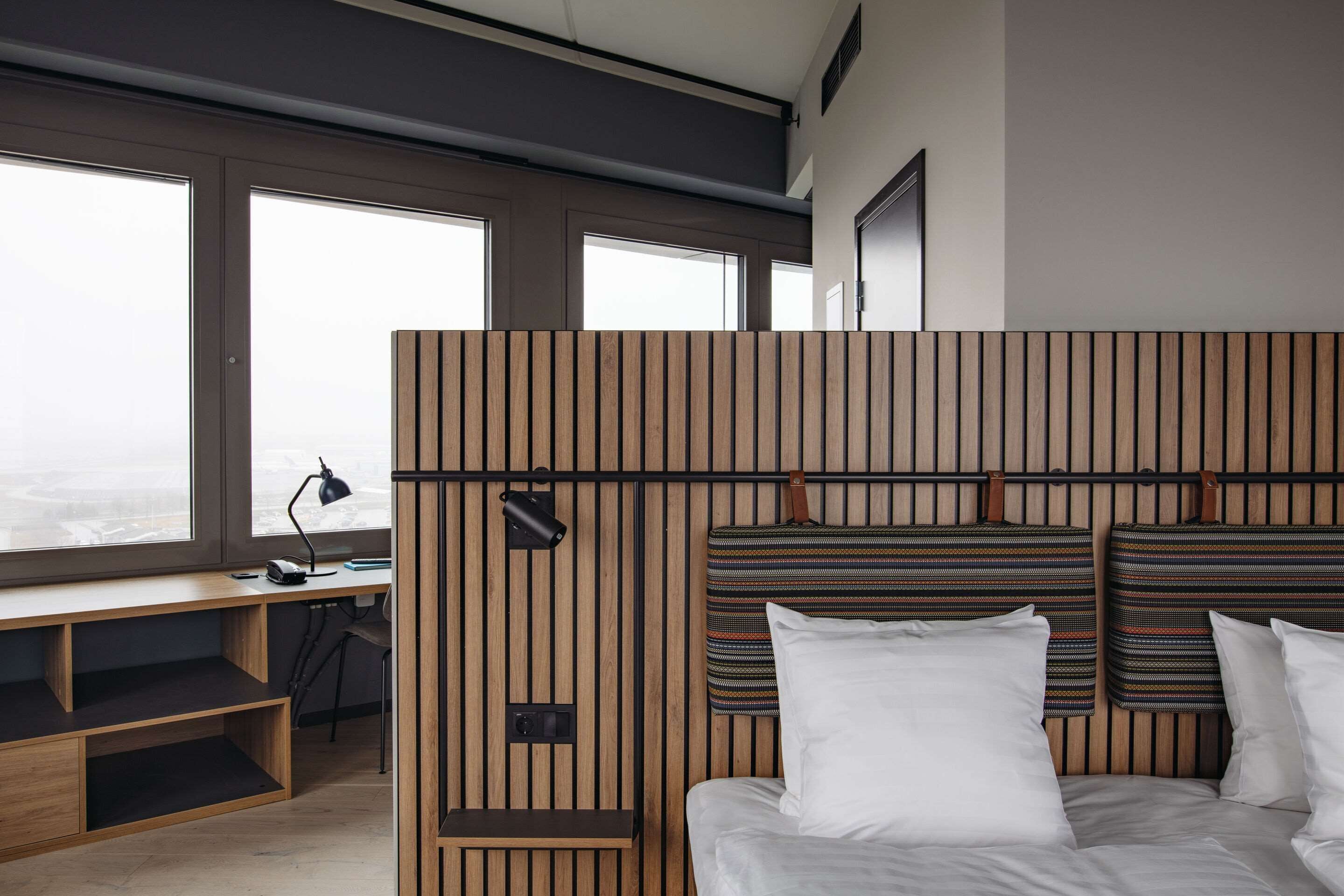 Habitación de hotel moderna con un ventanal grande, cabecera de madera, escritorio y cama con ropa blanca.