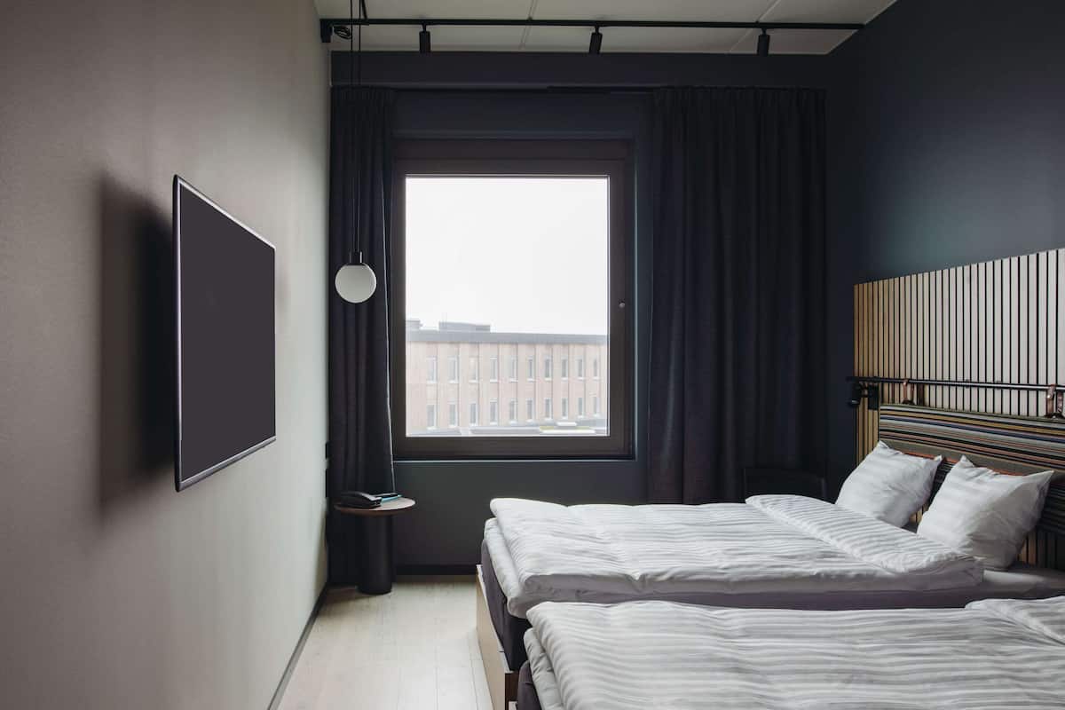 Una habitació d'hotel amb dos llits, una finestra gran amb cortines, un televisor fixat a la paret i una taula de nit amb una làmpada.