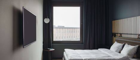 A hotel room with two beds, a large window with curtains, a wall-mounted TV, and a bedside table with a lamp.