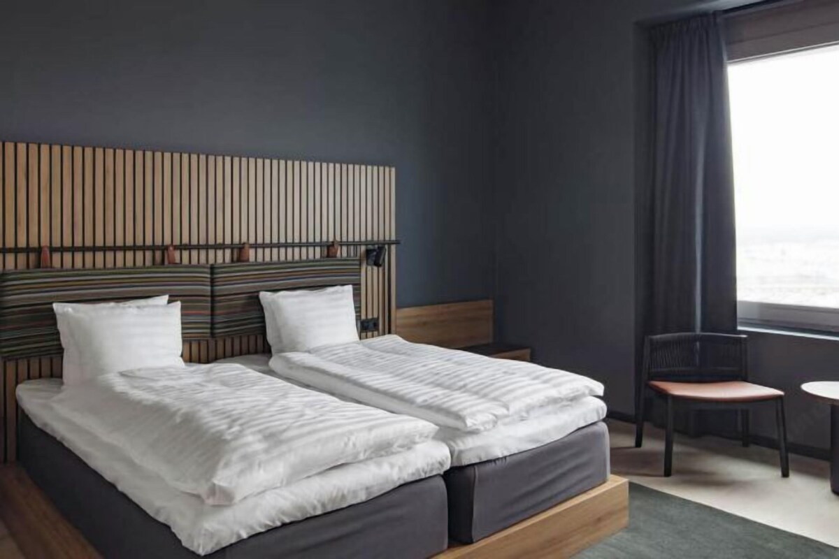 Habitación de hotel con cama, dos almohadas, cabecero, una silla, una mesita y una ventana con cortinas.