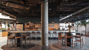 A modern bar area with a central round table surrounded by chairs, a counter with shelves of bottles, and a large pillar in the middle.