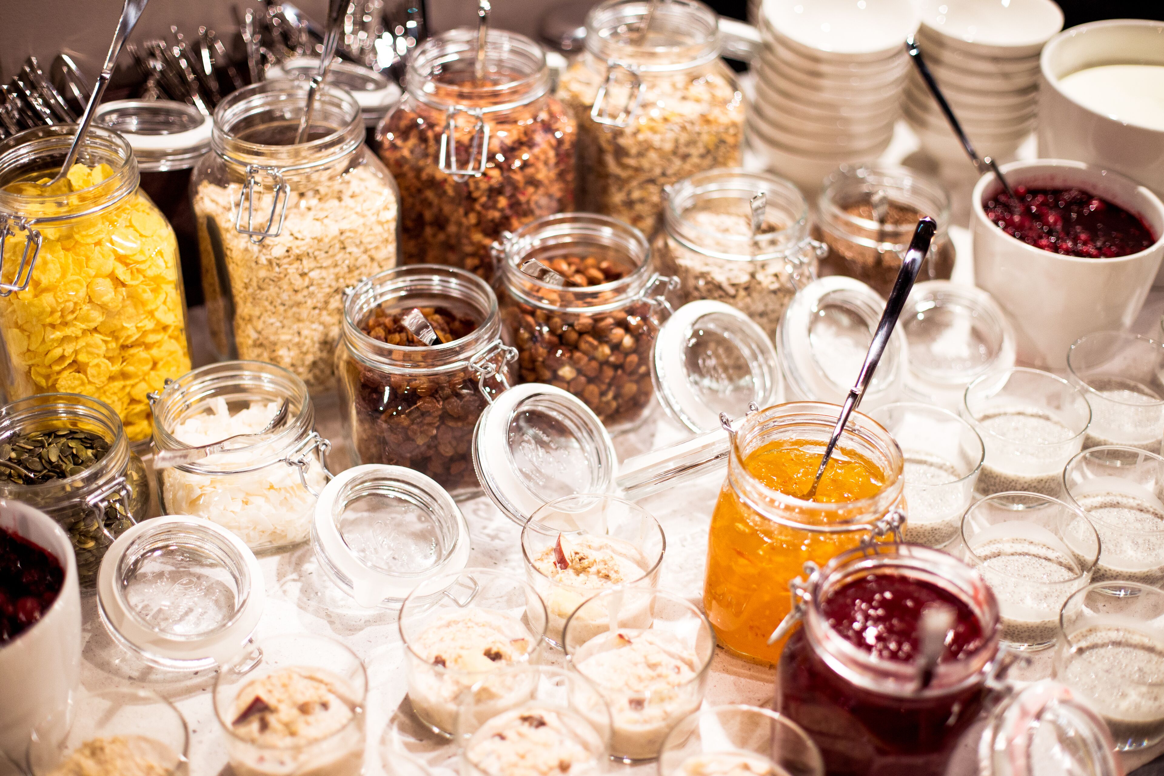 A variety of food items in jars and glasses, including cereals, nuts, and spreads, arranged on a table.