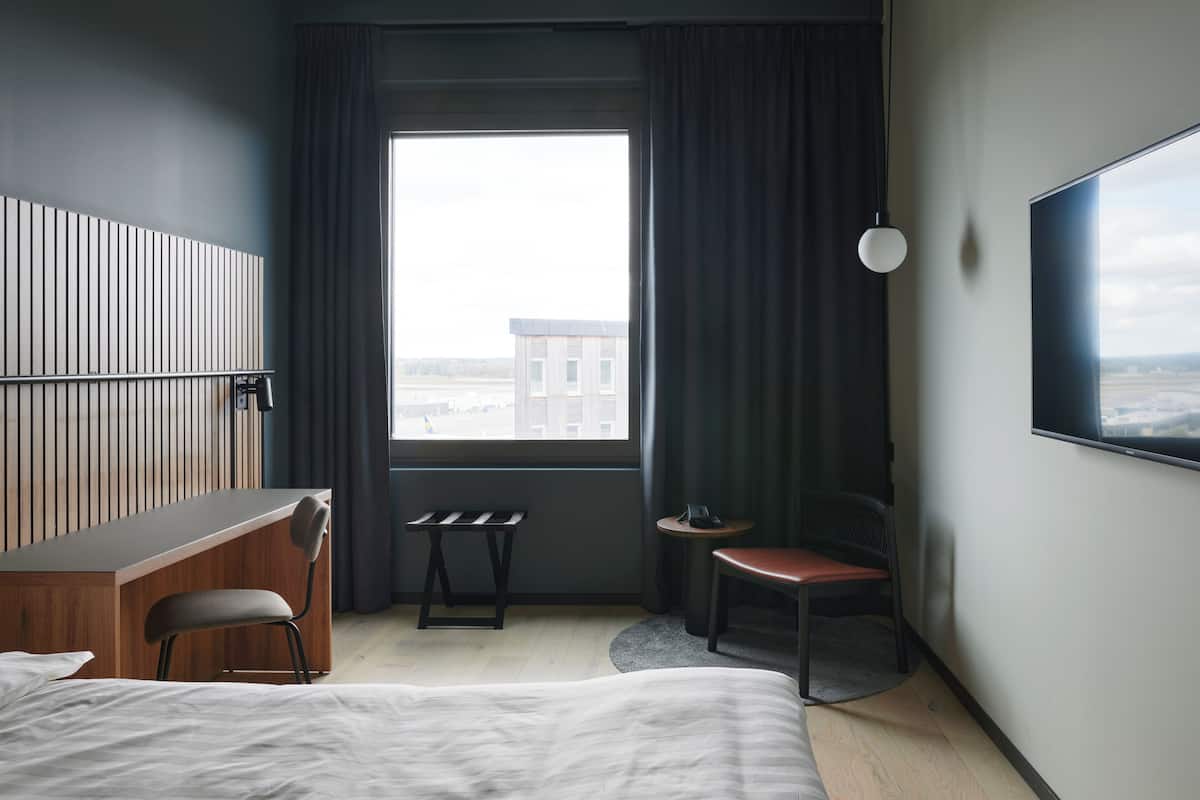 Habitació d'hotel amb llit, escriptori, cadira, taula petita, televisió i finestra amb cortines.