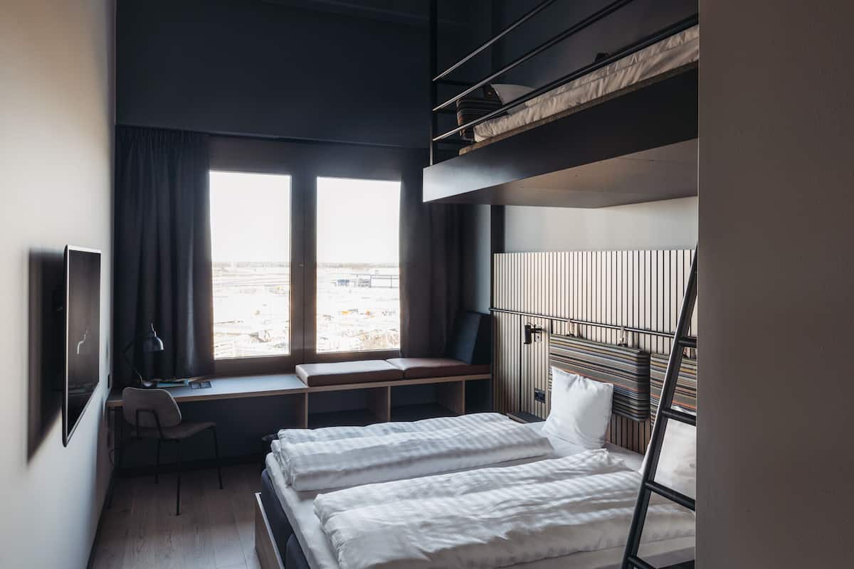 Una habitació d'hotel moderna amb una lliterola superposada, una taula de treball amb una cadira, una finestra amb cortines i vista exterior.