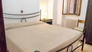 Ein Einzelbett mit gesteppter Matratze, ein Nachttisch mit Lampe, ein Heizkörper und ein Fenster mit Jalousien.