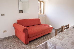 A room with a red sofa, a table with a patterned tablecloth, a window with blinds, and a radiator.