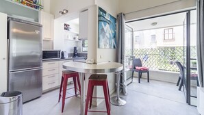 A modern kitchen with a stainless steel refrigerator, a microwave, and a white countertop. There are two red stools and a chair near the window. - Gordon Beach Ruppin street Best location (Tel Aviv-Yafo)