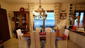 A dining room with a wooden table set for four, surrounded by white chairs with red trim. A chandelier hangs above, and a window offers a view of the sky and mountains.