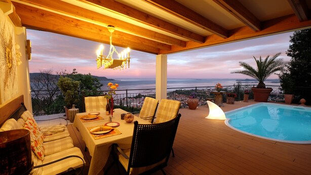 A wooden deck with a pool, outdoor seating, and a view of the sea at sunset.