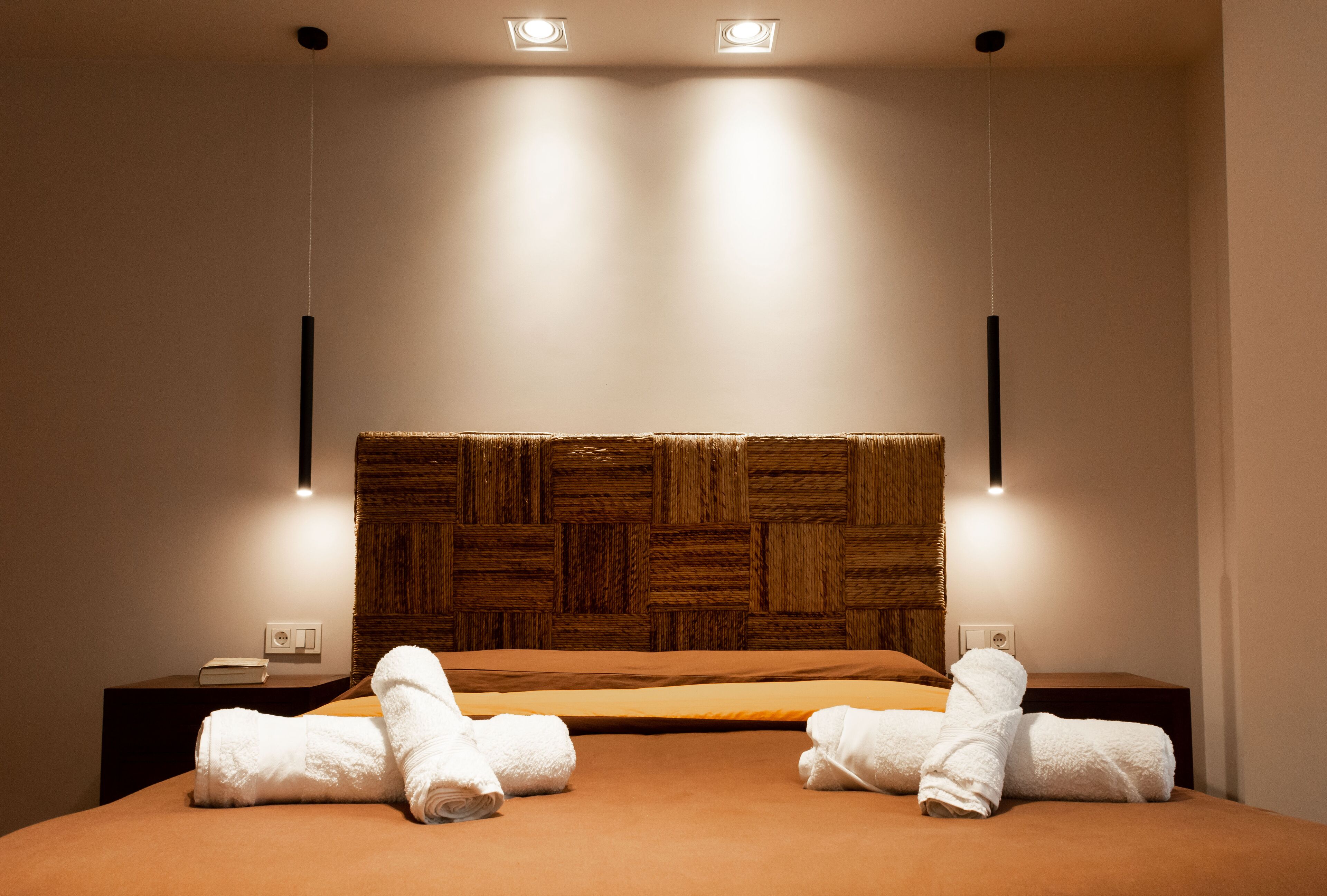 Une chambre d’hôtel avec une tête de lit en bois, deux tables de chevet, un lit recouvert d’une courtepointe marron et deux serviettes enroulées posées sur le lit.