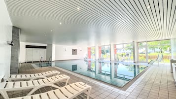 Ein moderner, überdachter Schwimmbereich mit großen Glaswänden, weißen Liegestühlen und Fliesenboden.
