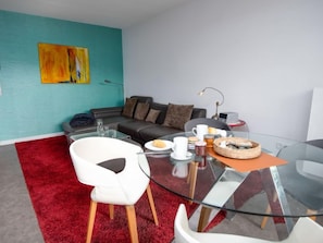 Ein modernes Wohnzimmer mit einem Glastisch, einem weißen Sessel, einem roten Teppich, einer Couch mit Kissen und einem Gemälde an der Wand.