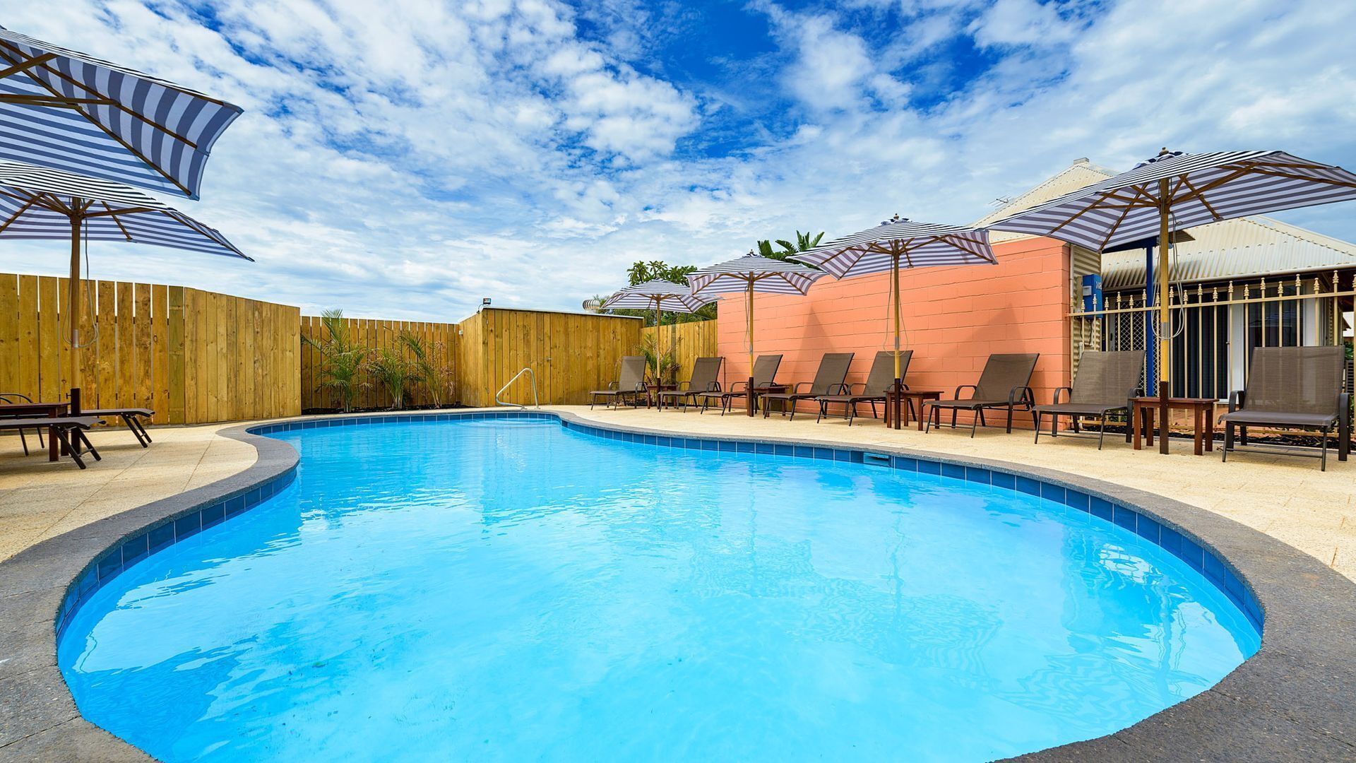 Piscina con sillas de descanso y sombrillas, rodeada por una valla de madera y un muro de ladrillo.