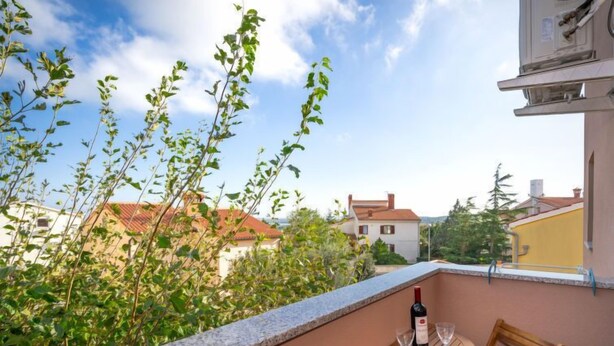A balcony with a table and chairs, a bottle of wine, and a view of buildings and greenery.