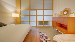 A bedroom with a bed, a wooden dresser, a nightstand, a lamp, a window with blinds, and a decorative mirror. - Leyteire large one bedroom apartment (BORDEAUX)