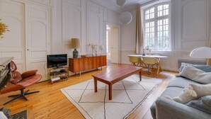 A living room with a wooden floor, a TV, a sofa, a coffee table, and a dining area with chairs and a table. - Leyteire large one bedroom apartment (BORDEAUX)