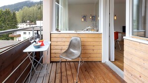 Ein Balkon mit Holzboden, einem Metallstuhl, einem kleinen Tisch mit Korb und einem Fenster mit Blick auf ein bewaldetes Gebiet.
