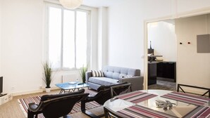 A modern living room with a sofa, coffee table, and chairs, a dining table with a striped table runner, and a large window with curtains. - Martinique Street (BORDEAUX)
