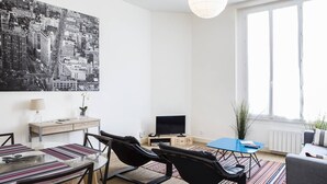 A modern living room with a cityscape painting, a flat-screen TV, a dining area with striped tablecloths, and a large window with a view. - Martinique Street (BORDEAUX)