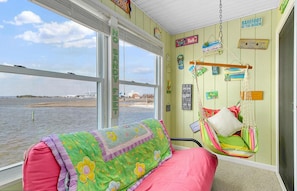 Una habitación con un amplio ventanal con vista a una masa de agua, un sillón colgante y una colcha de patchwork colorida.