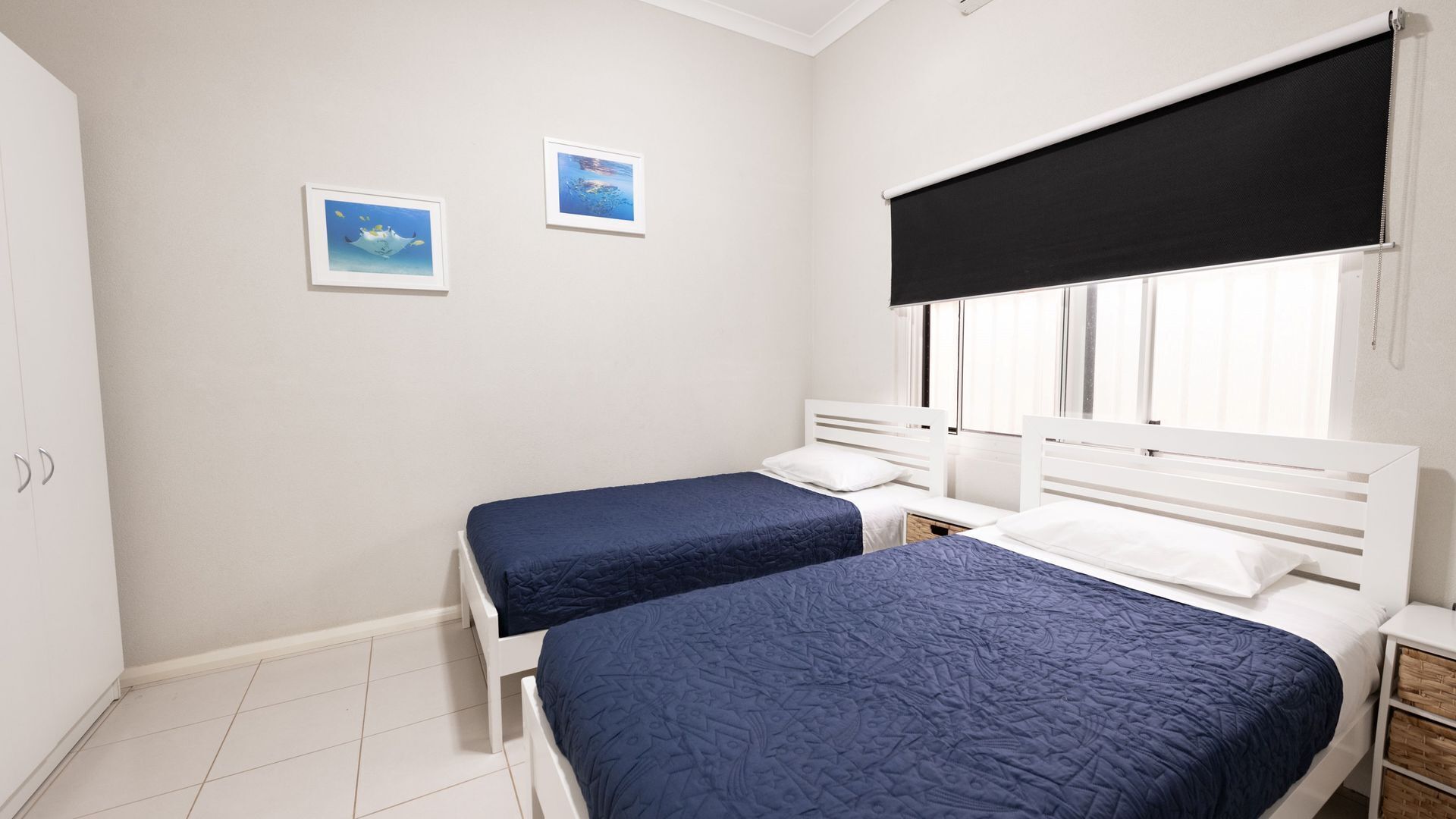 A bedroom with two single beds, white headboards, a large window with a black blind, and two framed pictures on the wall.