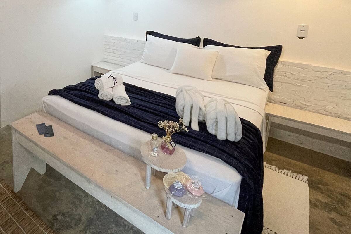A neatly made bed with white and dark blue bedding, a small bedside table with toiletries, and a bench at the foot of the bed.