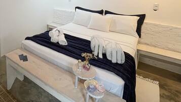 A neatly made bed with white and dark blue bedding, a small bedside table with toiletries, and a bench at the foot of the bed.