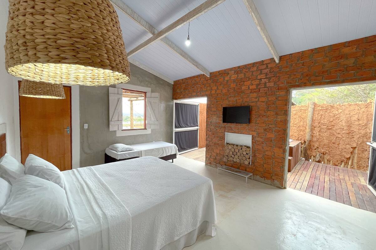 A room with a bed, a hanging light fixture, a brick wall, a television, and a view of the outside.