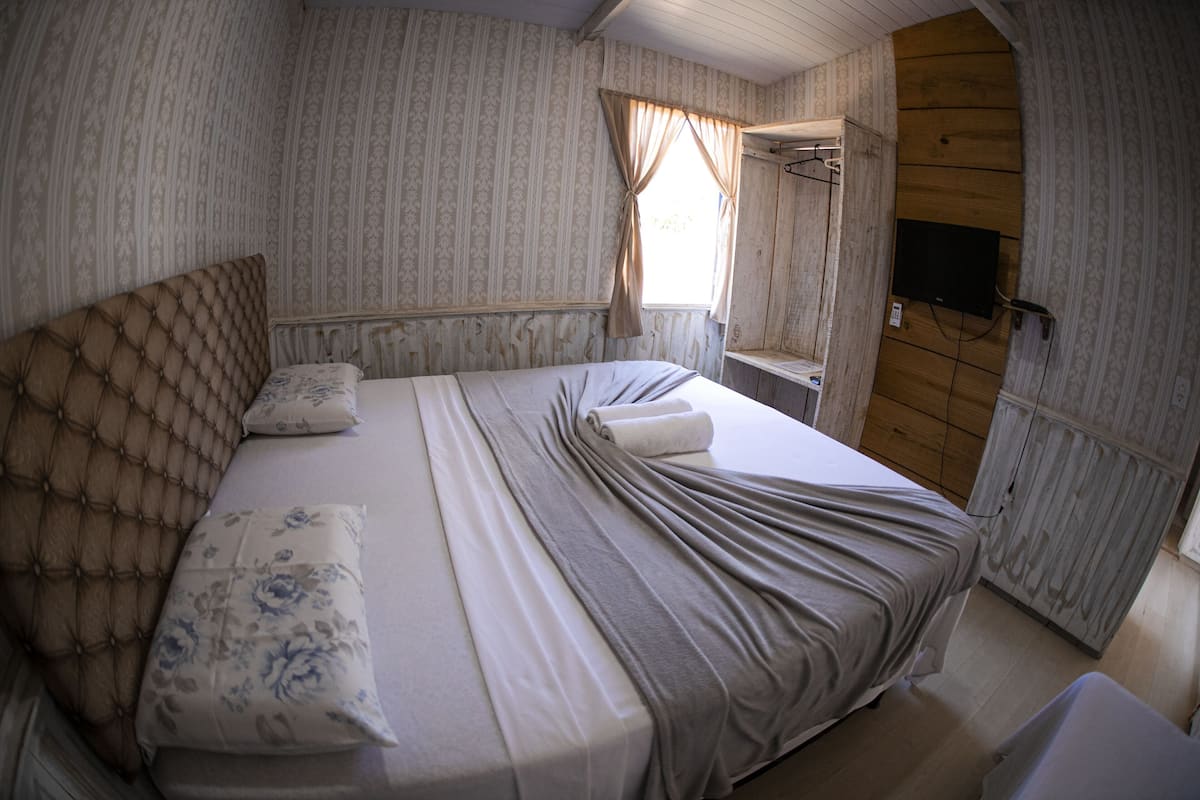 A neatly made bed with a headboard, a window with curtains, a television mounted on the wall, and a wooden wardrobe.