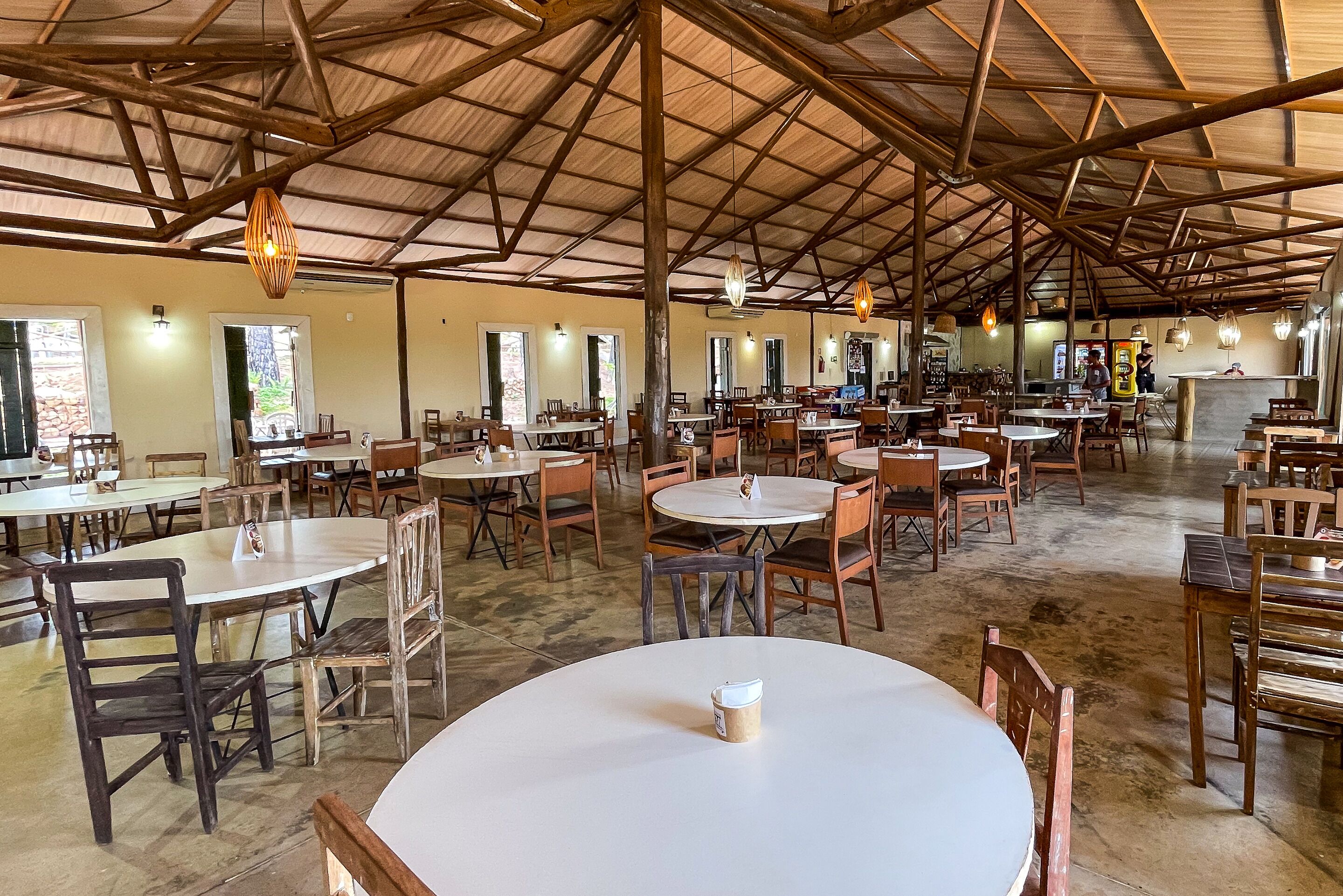 A spacious indoor dining area with multiple round tables and chairs, wooden beams on the ceiling, and natural light from windows.