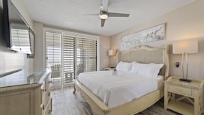 Une chambre à coucher comprenant un lit, des tables de chevet, un ventilateur de plafond, une fenêtre avec des stores et un téléviseur fixé au mur.
