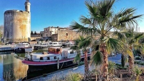 Une ville côtière dotée d’une imposante tour cylindrique, d’un port de plaisance avec des bateaux et d’un palmier au premier plan.