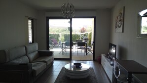 Un salon comprenant un canapé, une table basse, un téléviseur et une porte-fenêtre coulissante donnant sur une terrasse extérieure avec des chaises et une table.