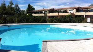 Une piscine dotée d’une terrasse en bois, entourée de verdure et de bâtiments avec des balcons.