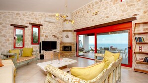 A spacious living room with stone walls, a fireplace, a television, and a view of the sea through large glass doors. - Sea View Nisaki Villa with private pool! (Zakynthos)
