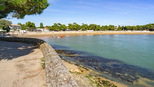 A coastal beach scene with a stone wall, clear water, and a row of buildings in the background. - La Grande Côte by Interhome (Saint Palais sur mer)