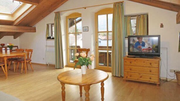 A cozy living room with a wooden dining table, a television, and a large window with curtains. - Wohnung 8 (Am Sonneneck) by Interhome (Ruhpolding)