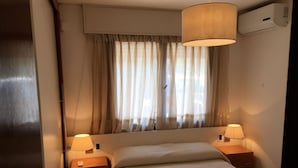 Una habitación de hotel con una cama, dos lámparas de noche, un ventanal con cortinas traslúcidas y un equipo de aire acondicionado en la pared.