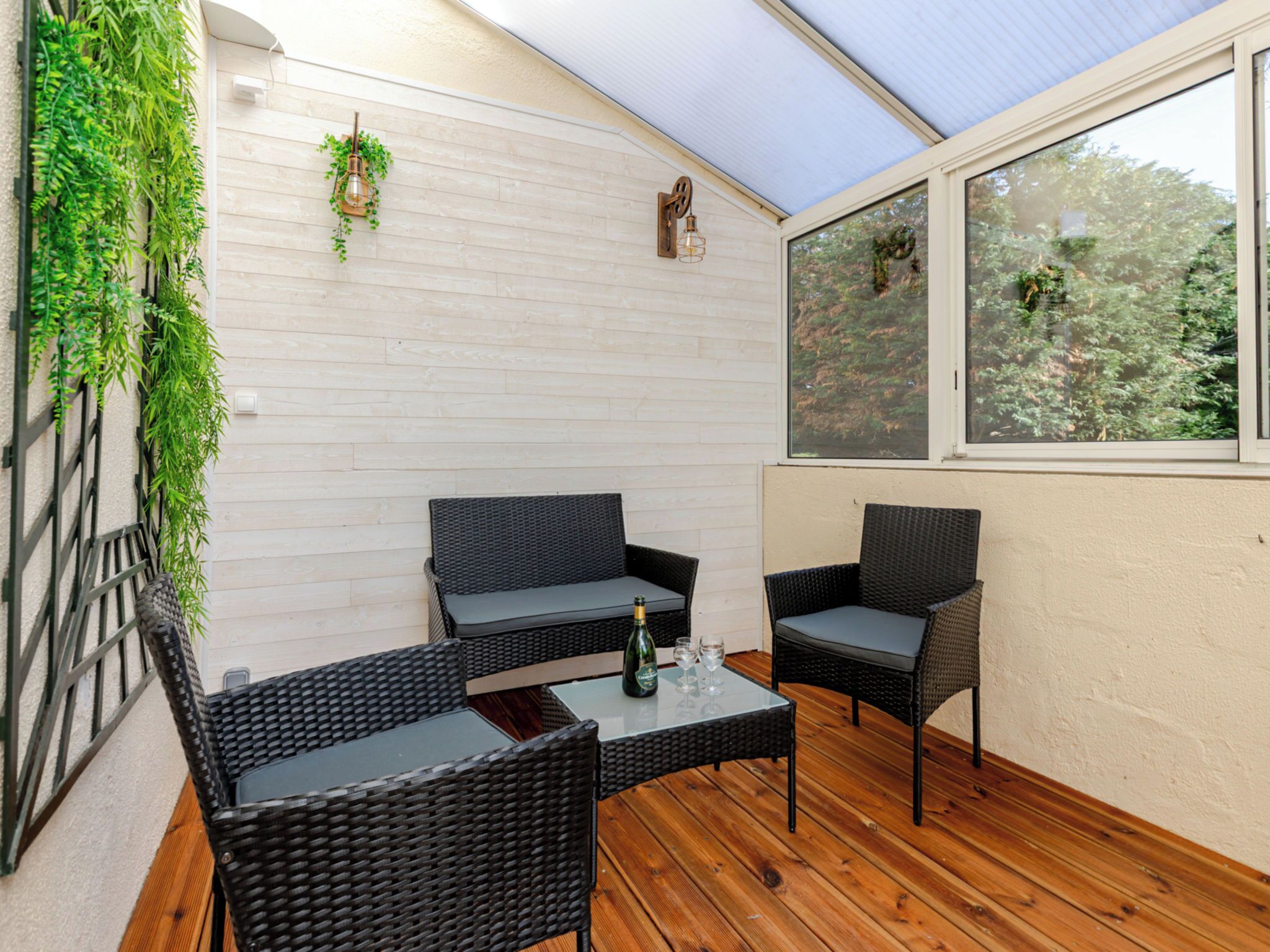 A covered patio with wicker furniture, a glass table, and a view of trees through the windows.
