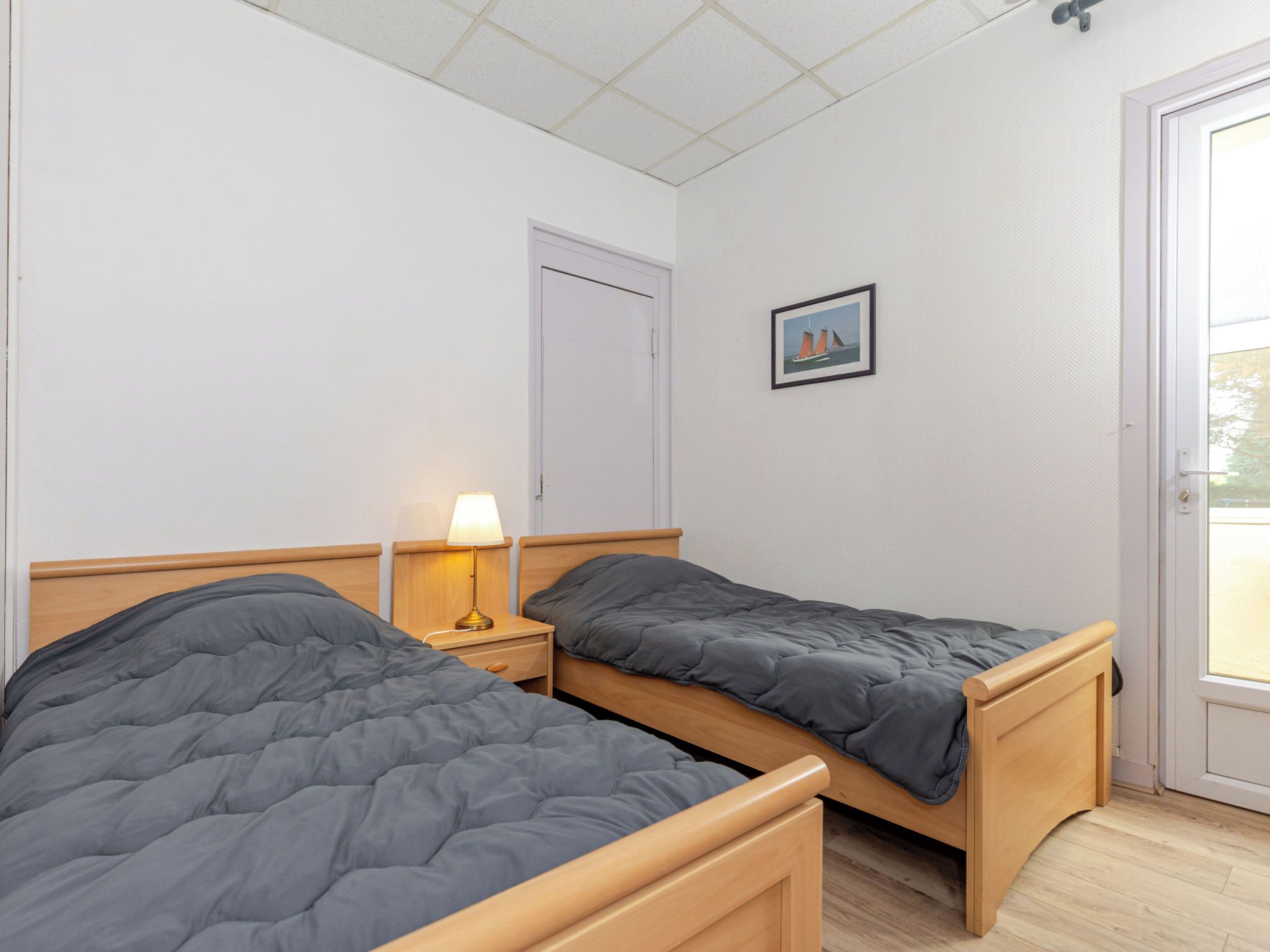 Ein Zimmer mit zwei Einzelbetten, einem Nachttisch mit Lampe, einer Tür und einem gerahmten Bild an der Wand.