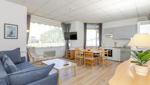 A modern living room with a sofa, a dining table with chairs, a TV mounted on the wall, and a kitchen area with cabinets and appliances. - Le Petit Robinson by Interhome (Dinard)