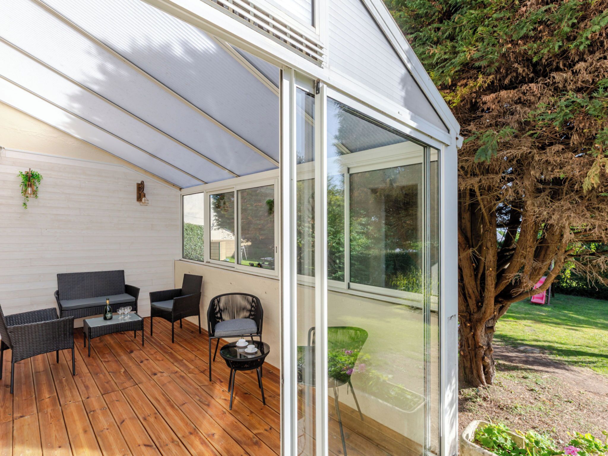 Une terrasse couverte, meublée de meubles en osier, une porte vitrée donnant sur l’extérieur et un grand arbre en arrière-plan.