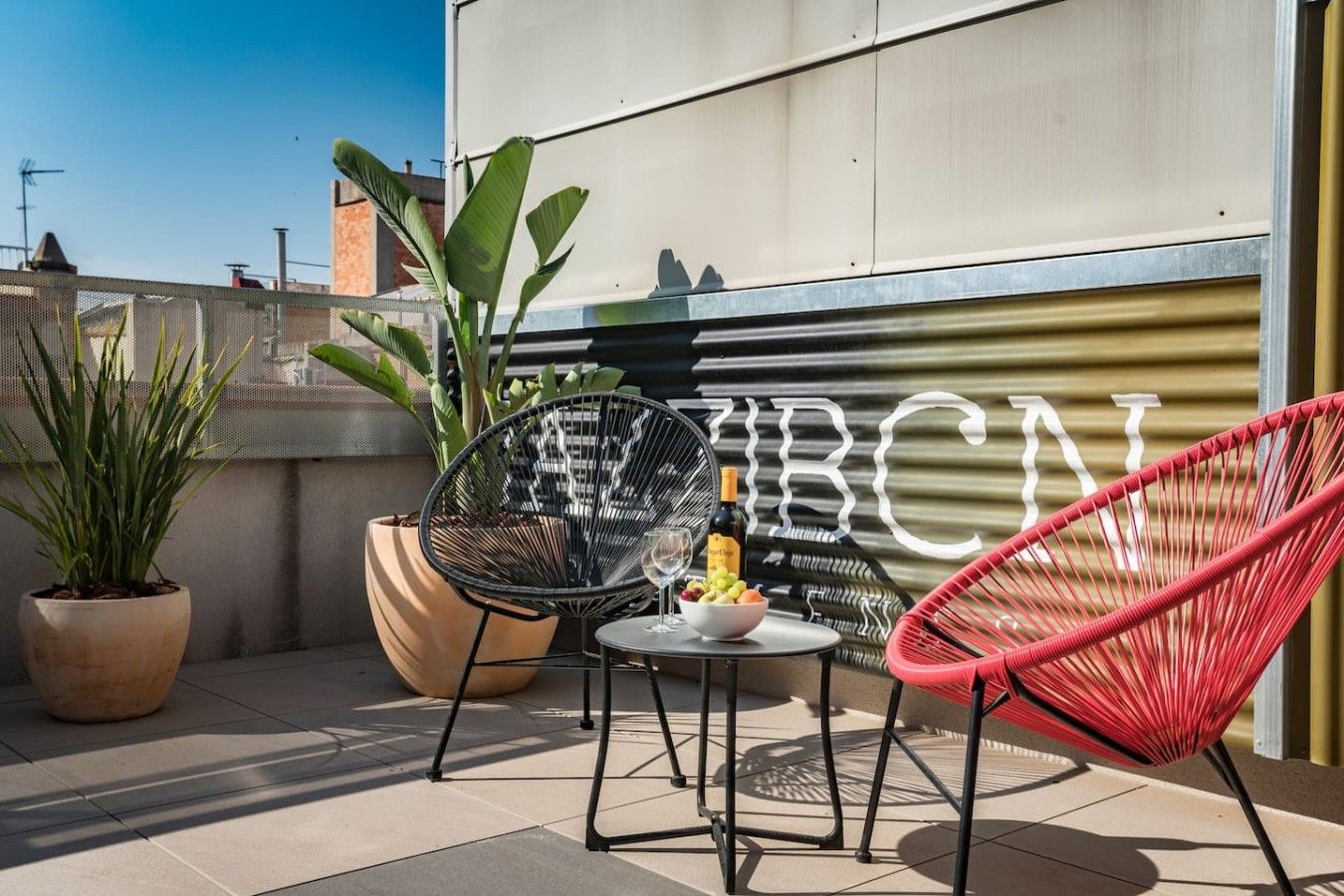 Une terrasse sur le toit avec deux chaises, une table sur laquelle se trouvent une bouteille et des fruits, ainsi que des plantes en pot.