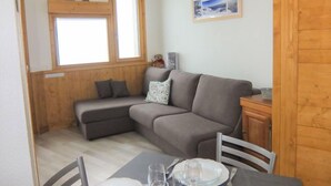 Een gezellige kamer met een grijze bank, een eettafel met stoelen en een raam met houten kozijnen.