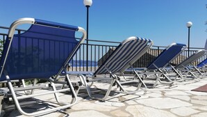 Una fila de tumbonas con estructura metálica y asientos de tela azul, dispuestas en un patio de baldosas de piedra con vista despejada al mar.