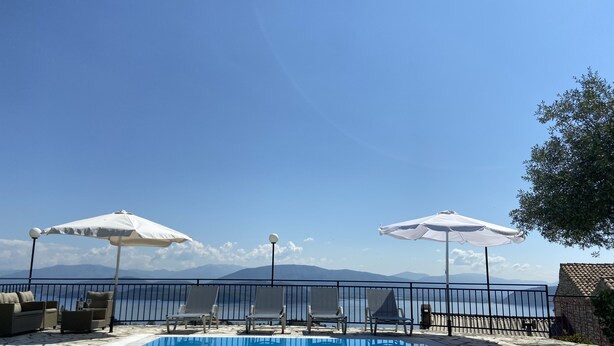 Una piscina exterior con tumbonas y sombrillas, rodeada por una terraza de piedra y vistas al mar y a las montañas.