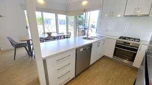 Eine moderne Küche mit weißen Schränken, einem Geschirrspüler, einem Herd und einem Spülbecken. Durch das Fenster ist eine Essecke mit Tisch und Stühlen zu sehen.